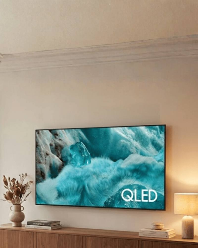 QLED TV Q7F mounted on a warm beige living room wall, displaying teal ocean waves above a wooden TV console with books, a vase, and a table lamp.
