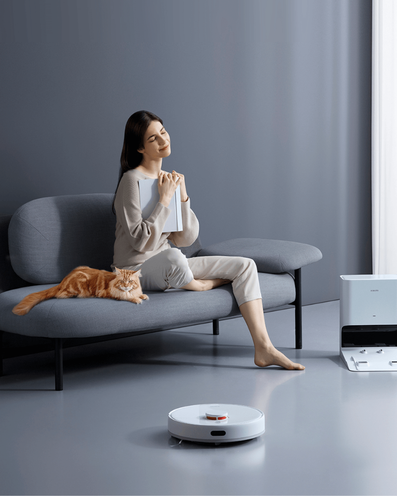 A promotional image for Xiaomi vacuum cleaners showing a woman relaxing on a sofa with a cat while a robot vacuum cleans the floor, conveying a clean and healthy home environment.
