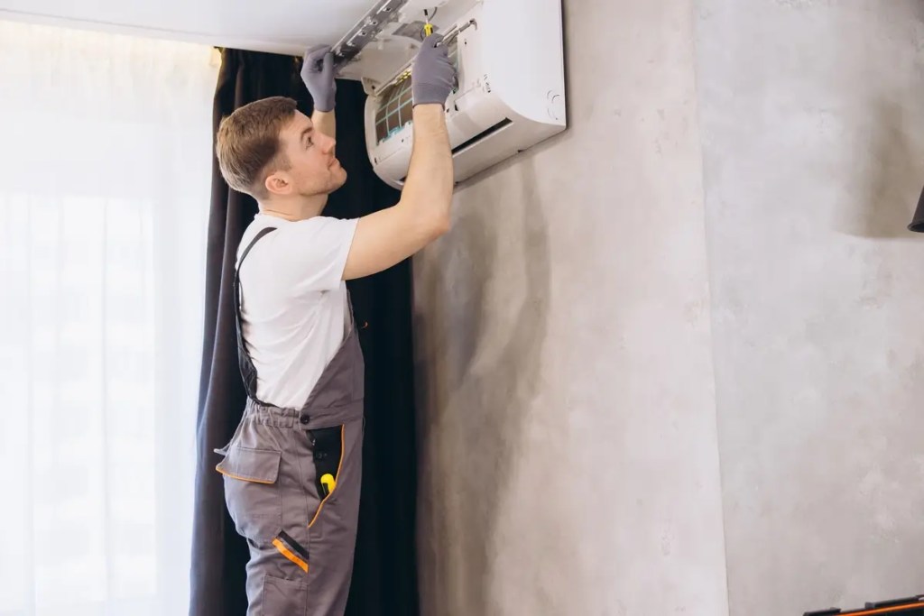 A professional technician in a blue uniform inspecting and repairing a wall-mounted air conditioner, highlighting the importance of regular aircond service and expert installation for inverter units.